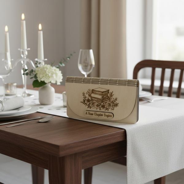 Holz-Umschlag aus hellem Birkensperrholz mit gelaserter Gravur A New Chapter Begins. Der Umschlag liegt dekorativ arrangiert, die natürliche Maserung und feinen Lasercut‑Details sind gut sichtbar. Ideal als Geschenkverpackung für Geld oder Gutscheine zu S